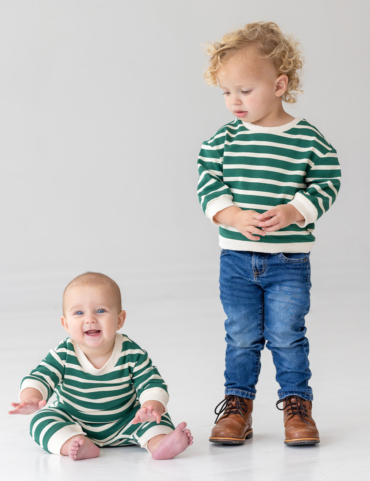 A baby smiles brightly while sitting barefoot in the Beckett and Bear Jackson Romper, next to a toddler in jeans and brown boots. Both are on a white floor, dressed in green and white stripes.