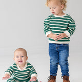 A baby smiles brightly while sitting barefoot in the Beckett and Bear Jackson Romper, next to a toddler in jeans and brown boots. Both are on a white floor, dressed in green and white stripes.