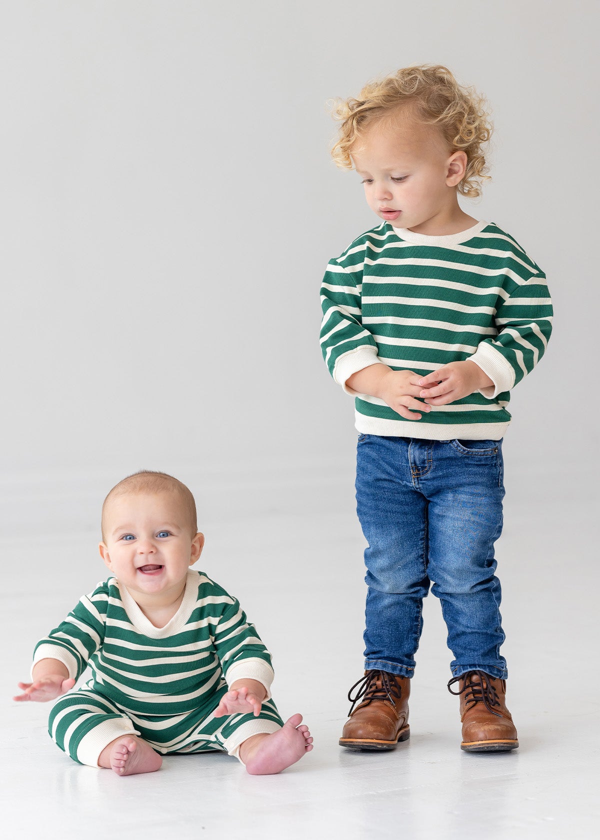 A baby smiles brightly while sitting barefoot in the Beckett and Bear Jackson Romper, next to a toddler in jeans and brown boots. Both are on a white floor, dressed in green and white stripes.