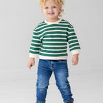 A young child with curly blonde hair wears the Beckett and Bear Jackson Top in green and white stripes, paired with blue jeans and brown lace-up boots, standing against a plain light gray background.