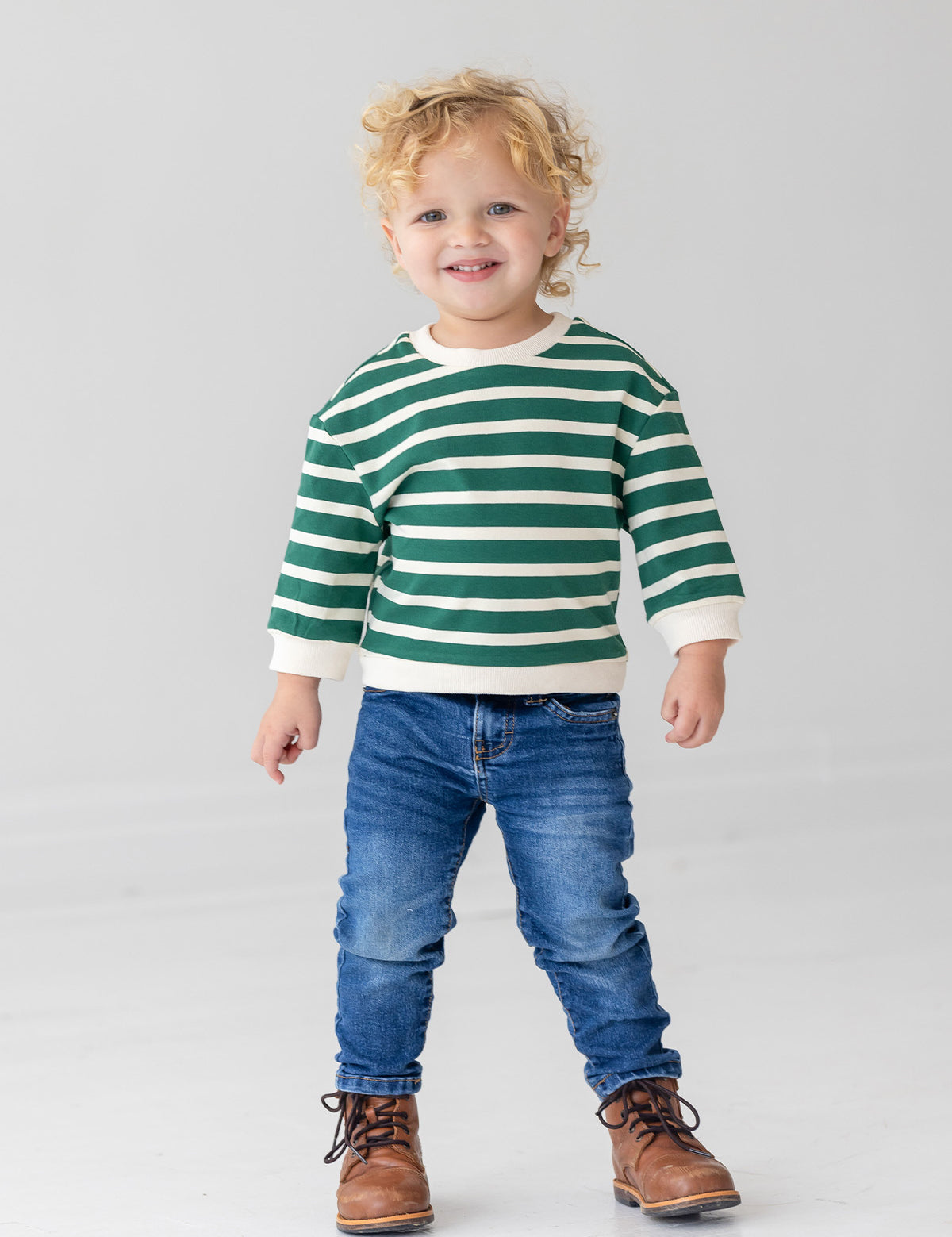 A young child with curly blonde hair wears the Beckett and Bear Jackson Top in green and white stripes, paired with blue jeans and brown lace-up boots, standing against a plain light gray background.