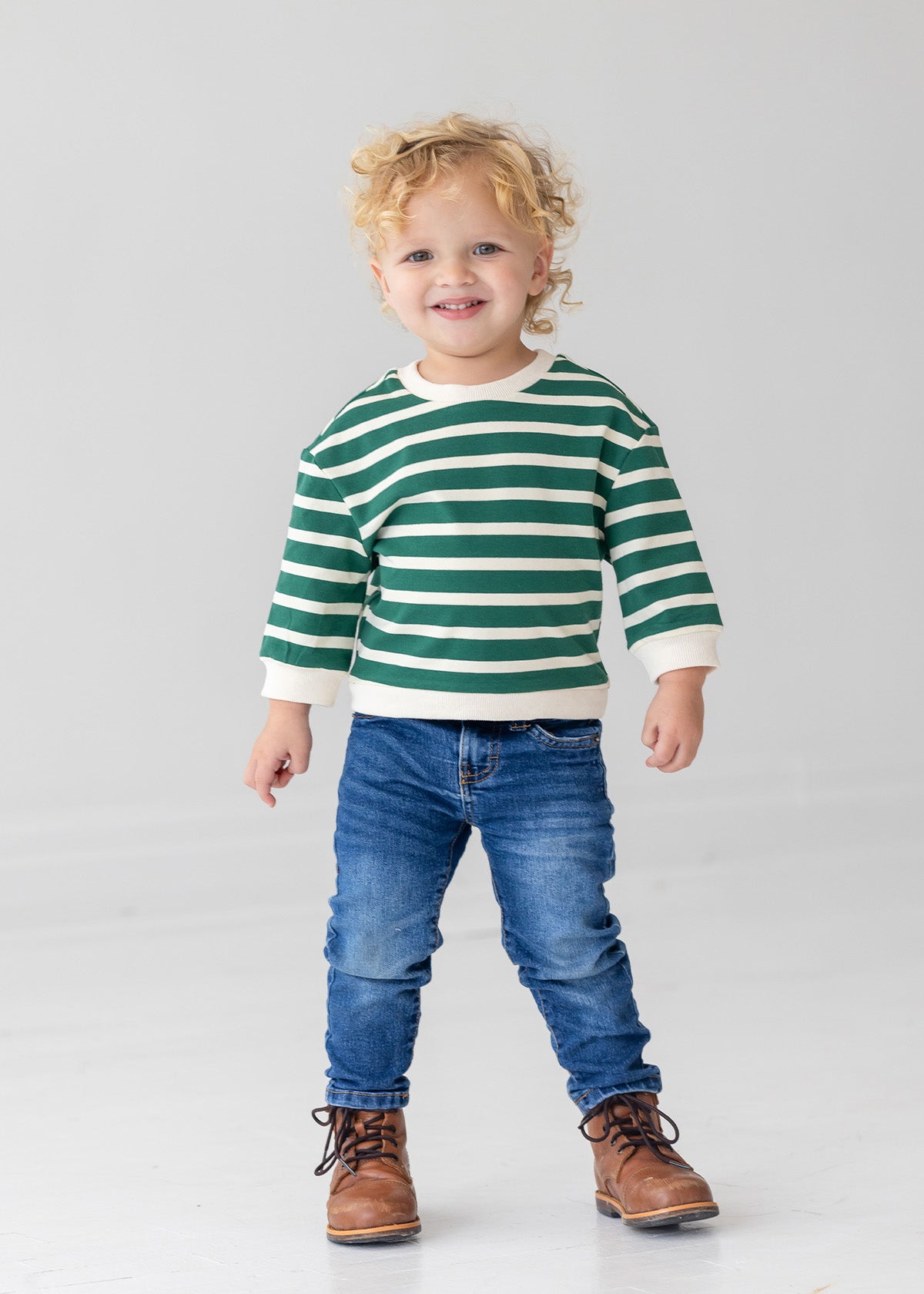 A young child with curly blonde hair wears the Beckett and Bear Jackson Top in green and white stripes, paired with blue jeans and brown lace-up boots, standing against a plain light gray background.