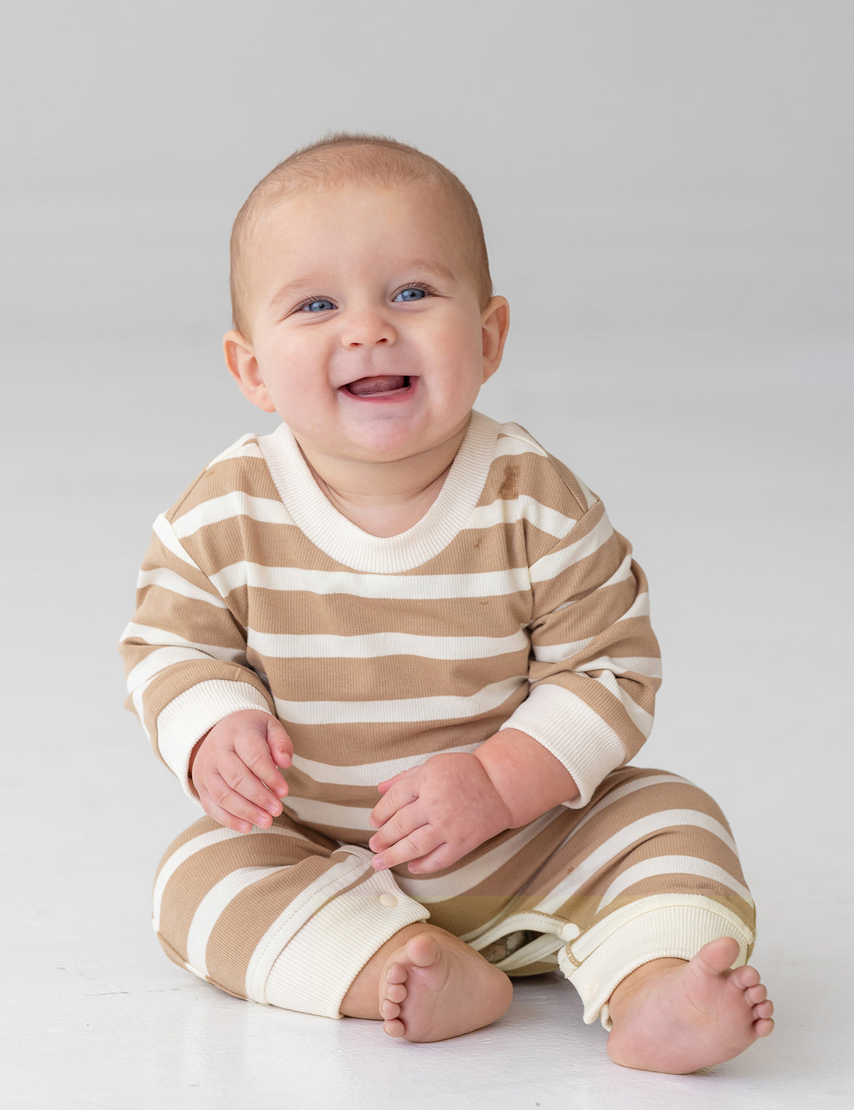 A smiling baby sits on the floor in the Beckett and Bear Mason Romper—beige and white stripes with cream cuffs—standing out against a plain, light background.