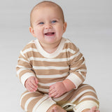A smiling baby sits on the floor in the Beckett and Bear Mason Romper—beige and white stripes with cream cuffs—standing out against a plain, light background.