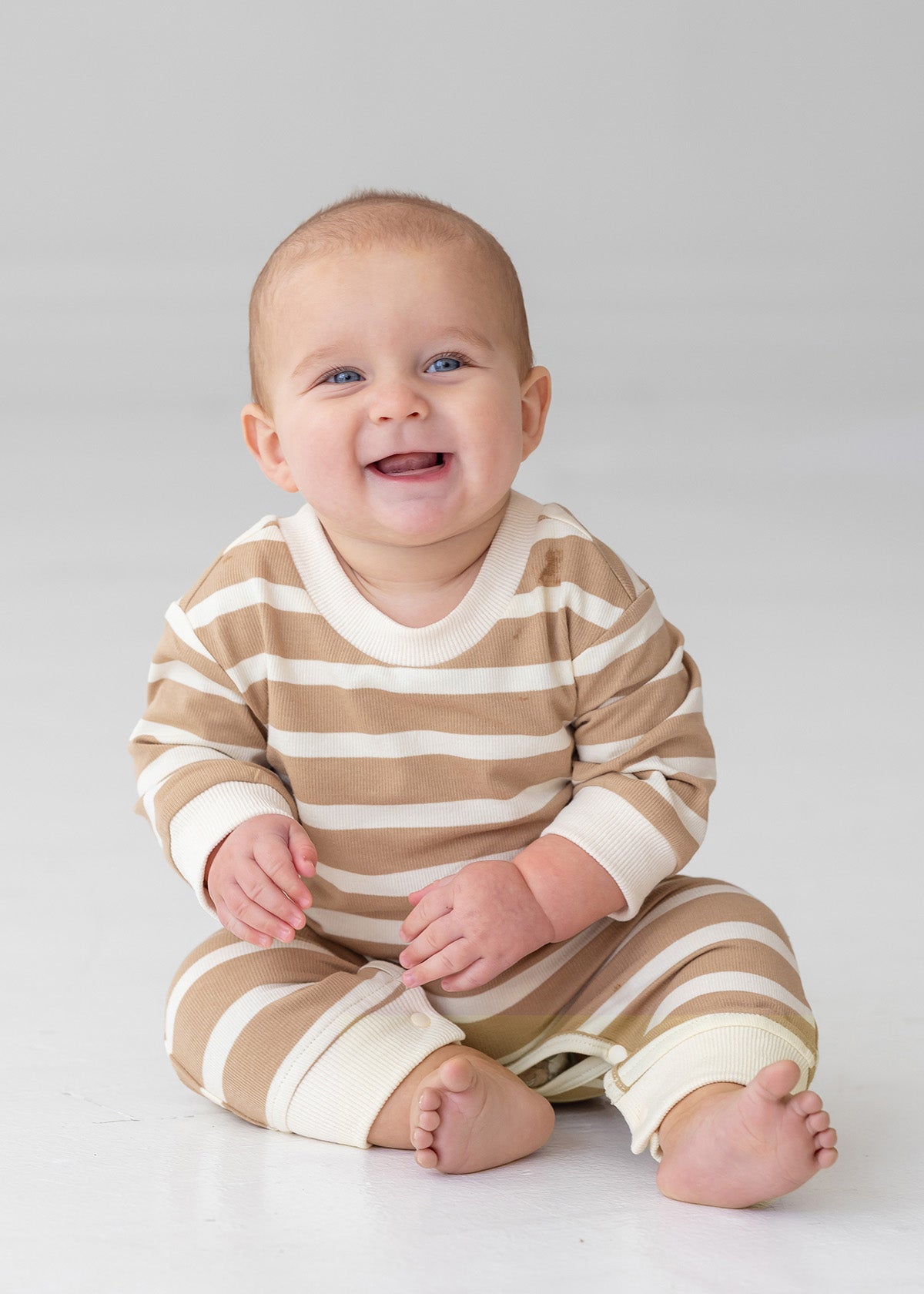 A smiling baby sits on the floor in the Beckett and Bear Mason Romper—beige and white stripes with cream cuffs—standing out against a plain, light background.