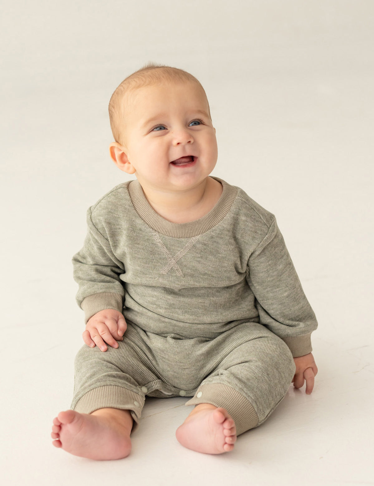 A smiling baby with light skin and short hair sits on a light-colored floor, wearing the Miles Romper by Beckett and Bear—a sage green, minimalist-inspired boys romper with ribbed fabric and matching pants.