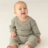 A smiling baby with light skin and short hair sits on a light-colored floor, wearing the Miles Romper by Beckett and Bear—a sage green, minimalist-inspired boys romper with ribbed fabric and matching pants.