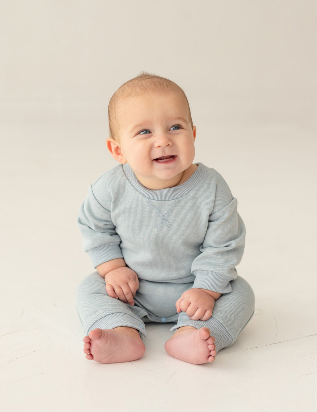 A baby with light hair and blue eyes smiles and looks to the side, sitting on the floor in the Beckett and Bear Aiden Romper against a simple, light background.