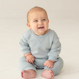 A baby with light hair and blue eyes smiles and looks to the side, sitting on the floor in the Beckett and Bear Aiden Romper against a simple, light background.