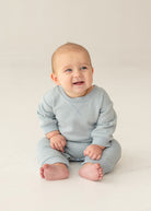 A baby with light hair and blue eyes smiles and looks to the side, sitting on the floor in the Beckett and Bear Aiden Romper against a simple, light background.