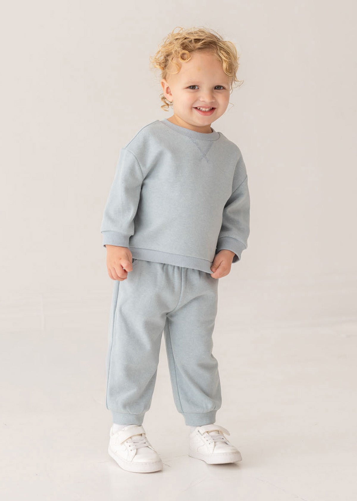 A young child with curly blonde hair smiles in the Beckett and Bear Aiden Two Piece Set—light blue sweatshirt and matching sweatpants with white sneakers—against a plain light background.