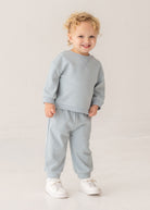 A young child with curly blonde hair smiles in the Beckett and Bear Aiden Two Piece Set—light blue sweatshirt and matching sweatpants with white sneakers—against a plain light background.