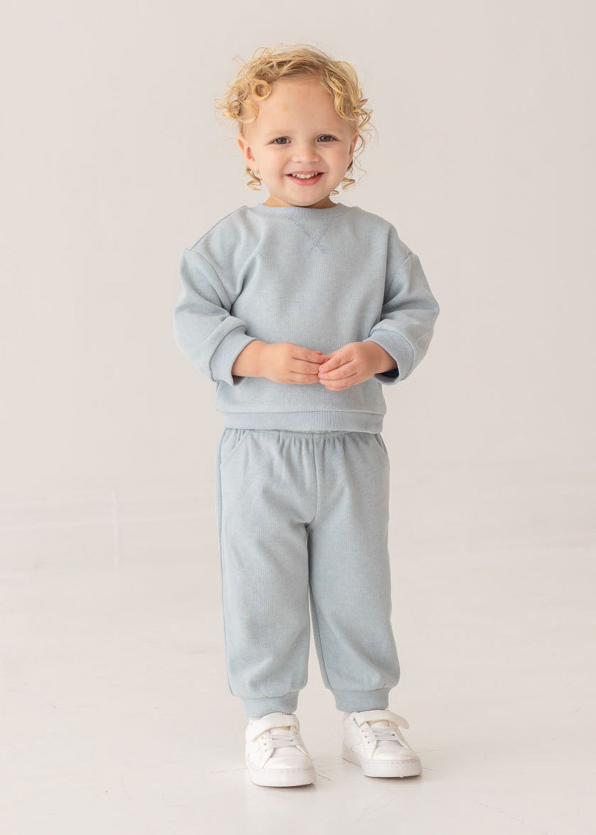 A smiling young child with curly blond hair stands against a plain background, wearing the stylish Beckett and Bear Aiden Two Piece Set—light blue sweatshirt and matching sweatpants—paired with crisp white sneakers.