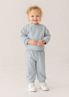 A smiling young child with curly blond hair stands against a plain background, wearing the stylish Beckett and Bear Aiden Two Piece Set—light blue sweatshirt and matching sweatpants—paired with crisp white sneakers.