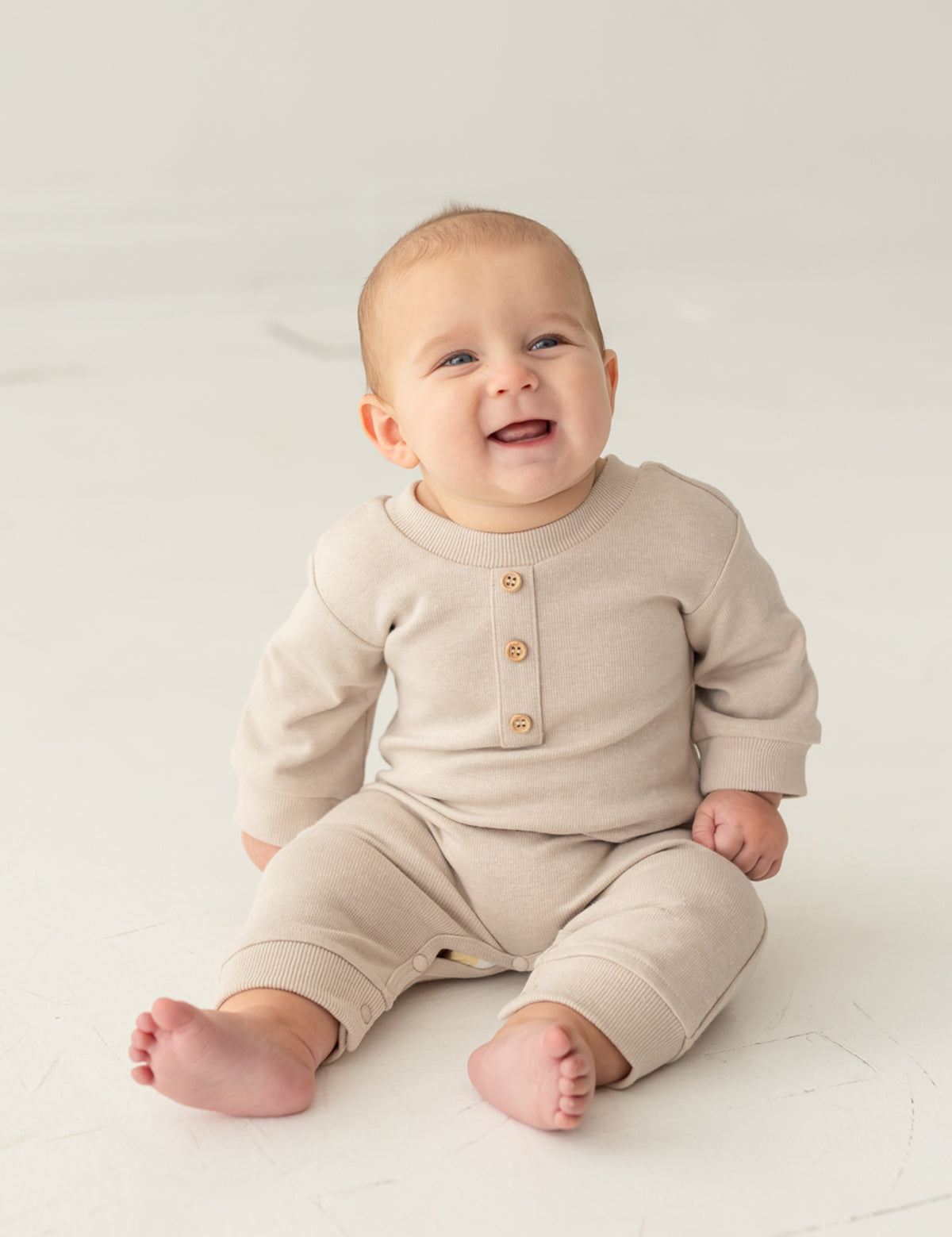 A smiling baby sits barefoot on a light floor against a neutral background, wearing the neutral-tone Isaac Romper by Beckett and Bear, featuring wooden buttons.