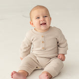 A smiling baby sits barefoot on a light floor against a neutral background, wearing the neutral-tone Isaac Romper by Beckett and Bear, featuring wooden buttons.