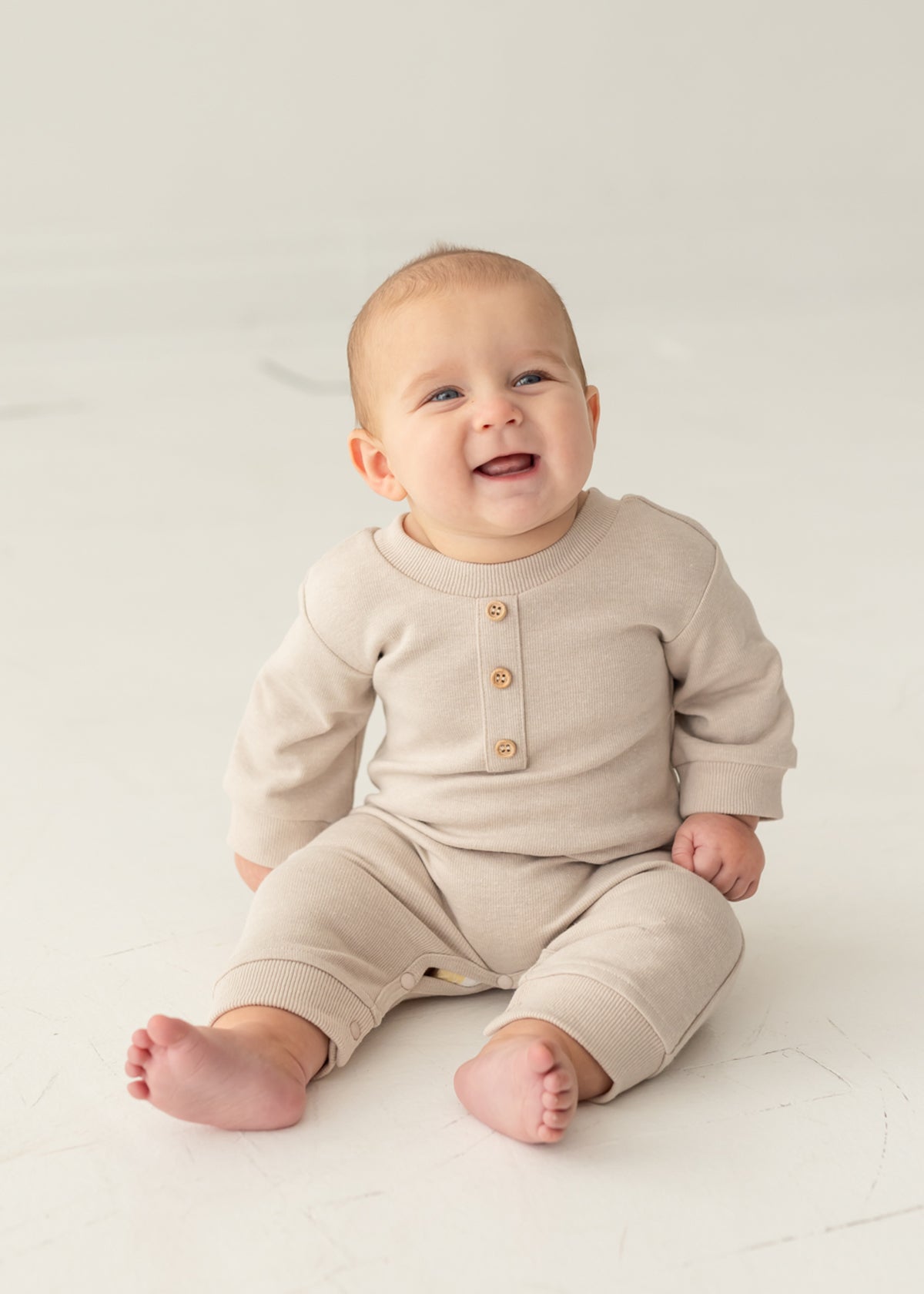 A smiling baby sits barefoot on a light floor against a neutral background, wearing the neutral-tone Isaac Romper by Beckett and Bear, featuring wooden buttons.