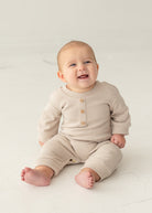 A smiling baby sits barefoot on a light floor against a neutral background, wearing the neutral-tone Isaac Romper by Beckett and Bear, featuring wooden buttons.