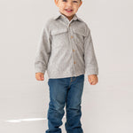 A young boy with short brown hair smiles while standing in a light gray Beckett and Bear Grayson Jacket over a button-up shirt, blue jeans, and tan shoes, set against a plain white background.