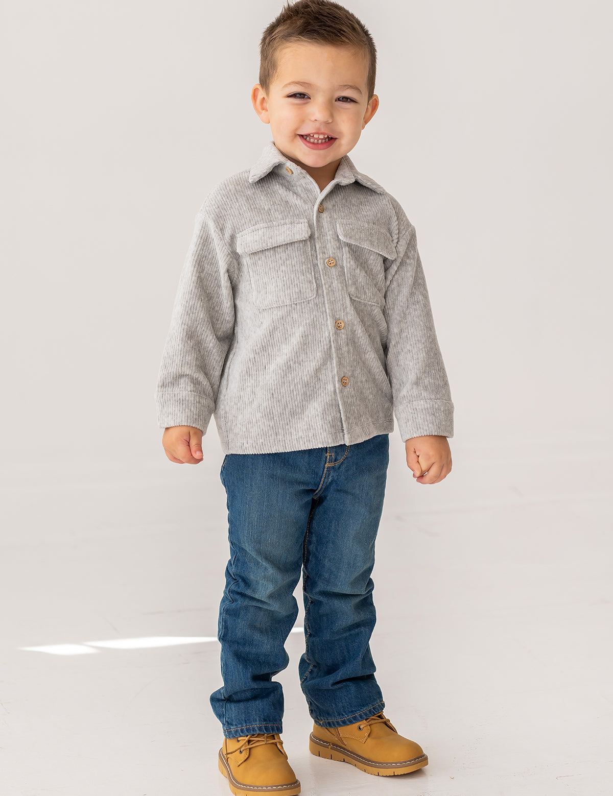 A young boy with short brown hair smiles while standing in a light gray Beckett and Bear Grayson Jacket over a button-up shirt, blue jeans, and tan shoes, set against a plain white background.