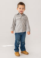A young boy with short brown hair smiles while standing in a light gray Beckett and Bear Grayson Jacket over a button-up shirt, blue jeans, and tan shoes, set against a plain white background.