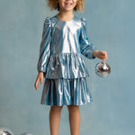 A young girl with curly hair, wearing the Isobella and Chloe Blue Stardust Dress and silver shoes, smiles while holding a small disco ball. Three more disco balls are on the floor beside her against a light blue background.