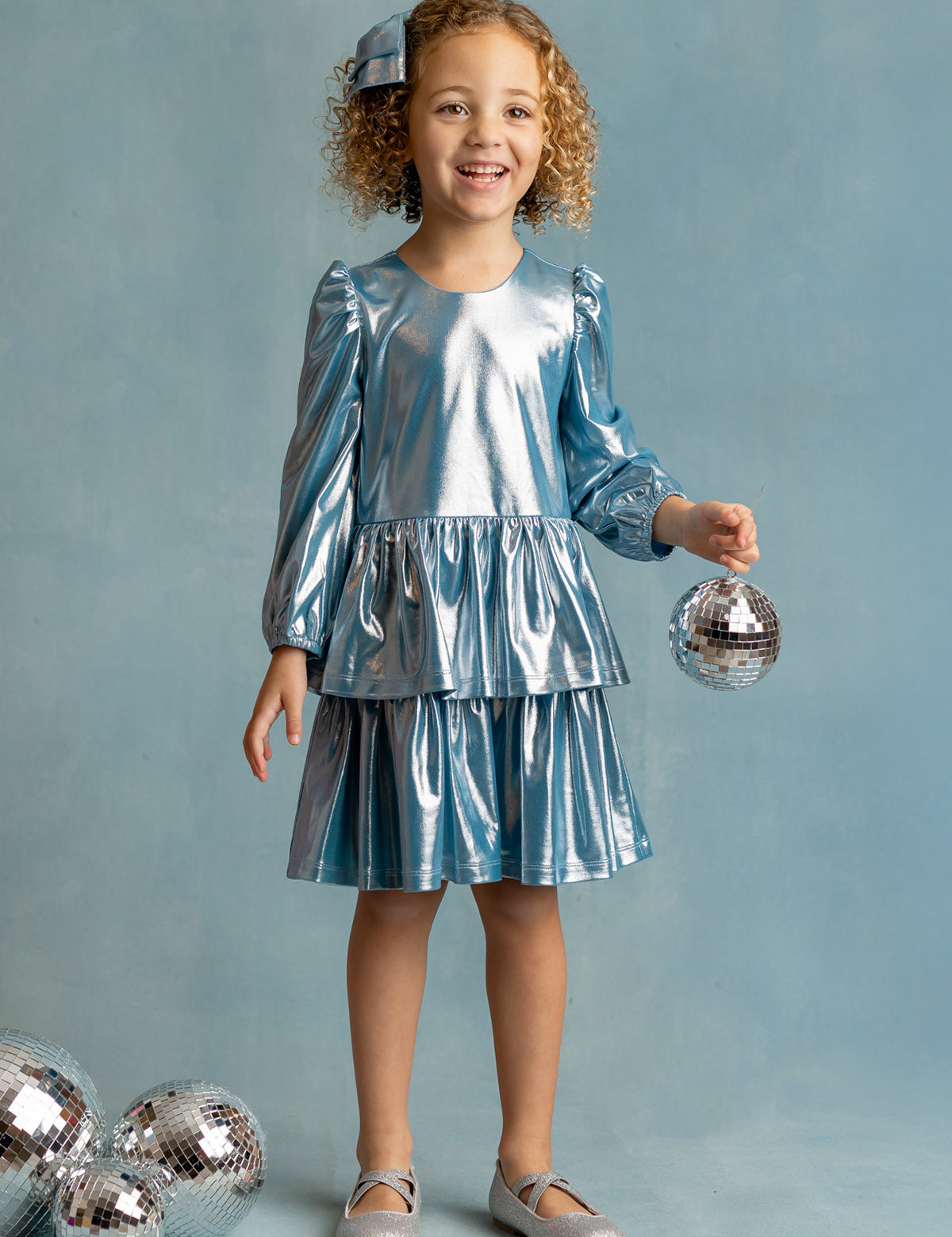 A young girl with curly hair, wearing the Isobella and Chloe Blue Stardust Dress and silver shoes, smiles while holding a small disco ball. Three more disco balls are on the floor beside her against a light blue background.