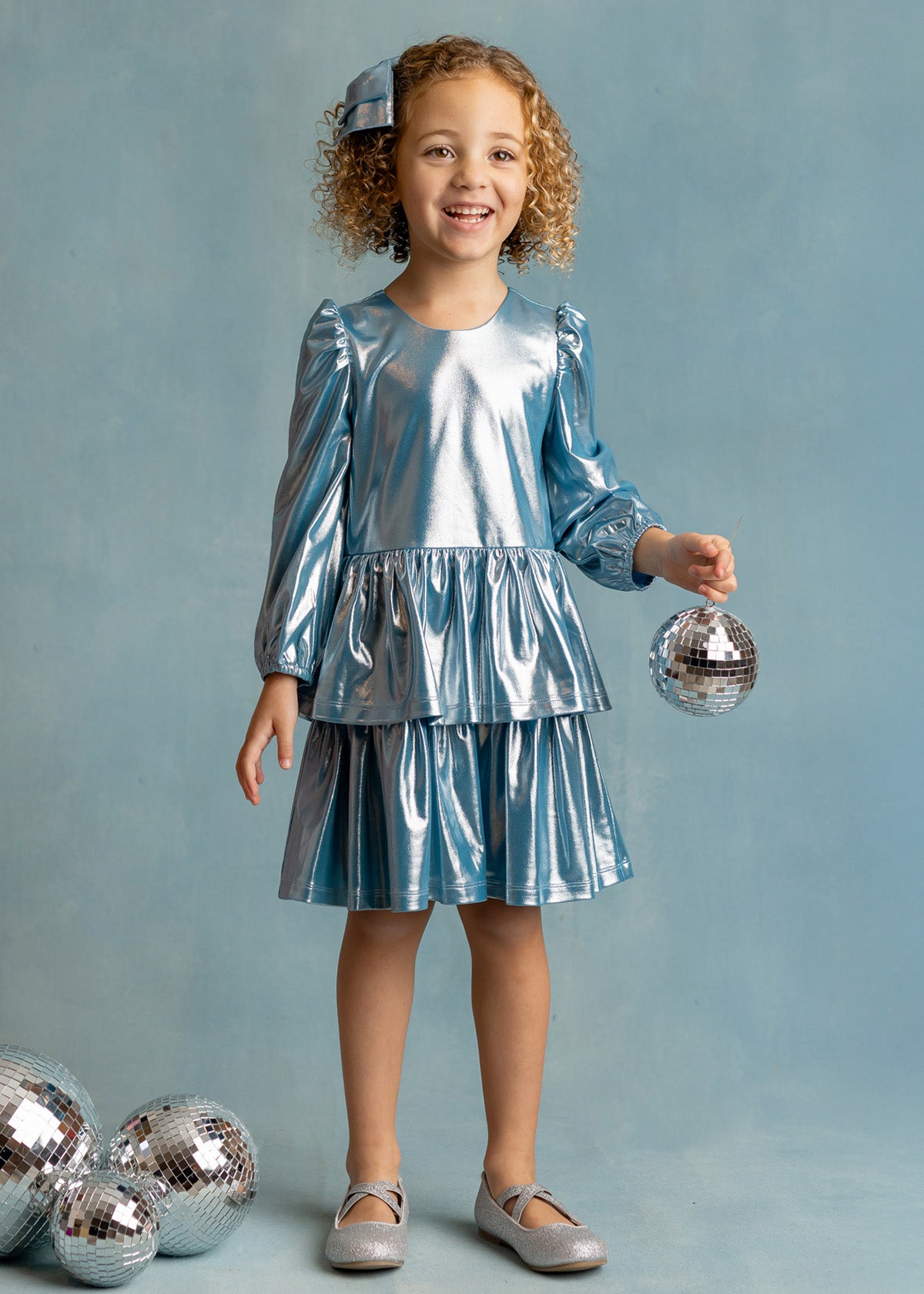 A young girl with curly hair, wearing the Isobella and Chloe Blue Stardust Dress and silver shoes, smiles while holding a small disco ball. Three more disco balls are on the floor beside her against a light blue background.