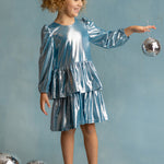 A young girl smiles in the Isobella and Chloe Blue Stardust Dress with a matching bow, balancing a disco ball in her hand. Three more disco balls rest at her feet against a pale blue background—perfect for special occasions.