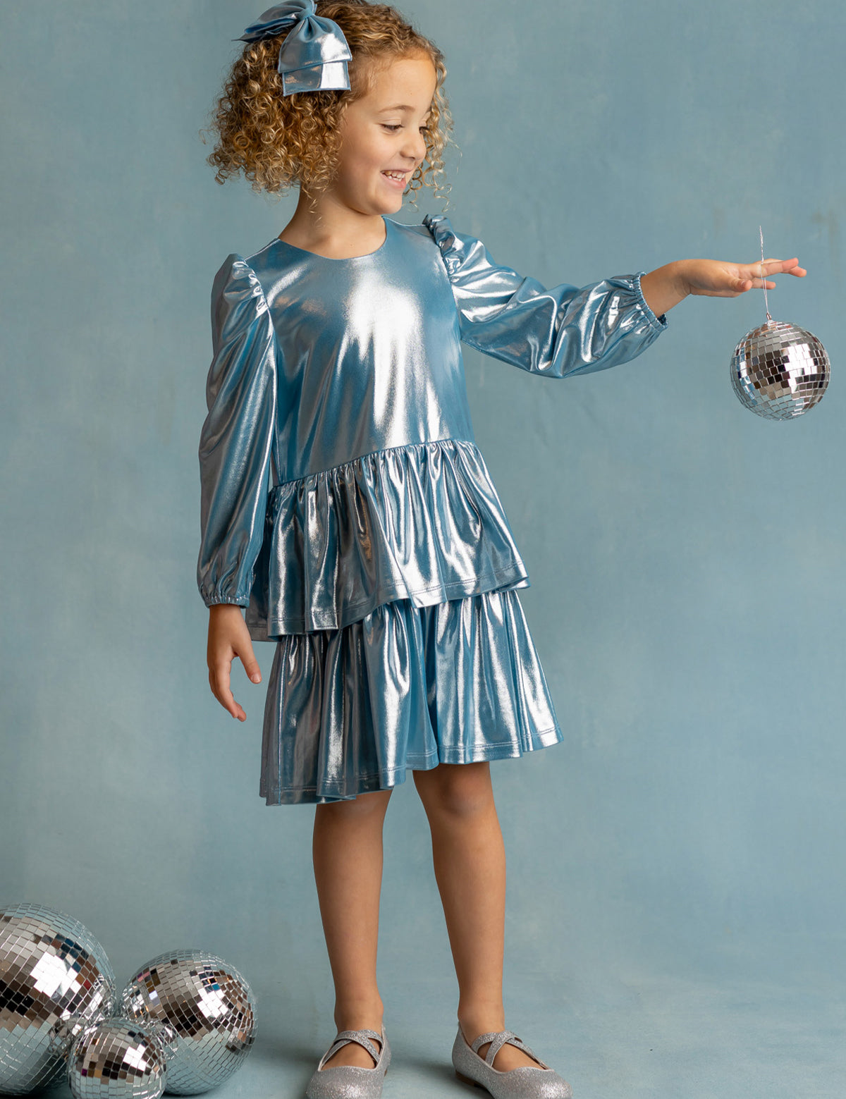 A young girl smiles in the Isobella and Chloe Blue Stardust Dress with a matching bow, balancing a disco ball in her hand. Three more disco balls rest at her feet against a pale blue background—perfect for special occasions.