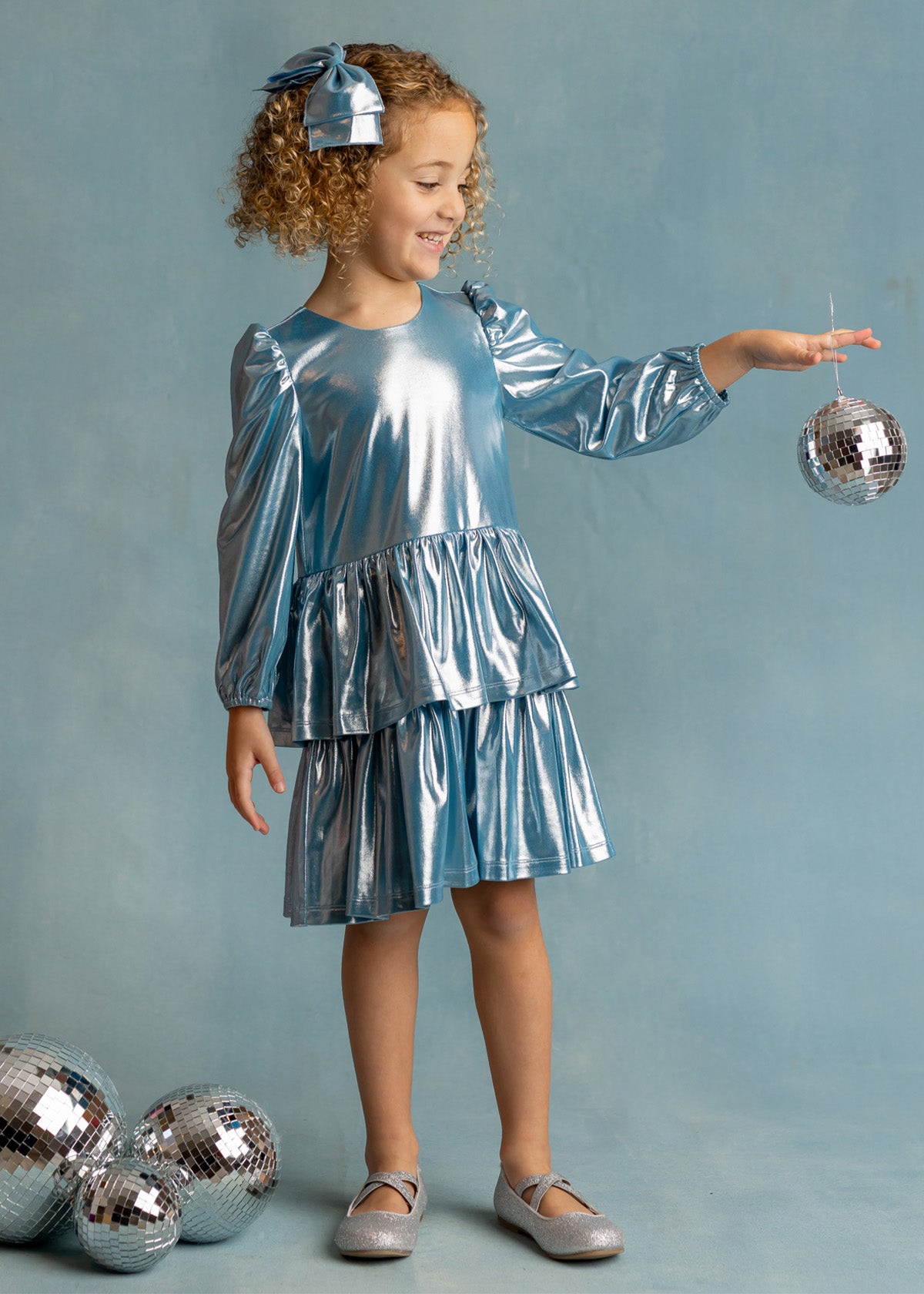 A young girl smiles in the Isobella and Chloe Blue Stardust Dress with a matching bow, balancing a disco ball in her hand. Three more disco balls rest at her feet against a pale blue background—perfect for special occasions.