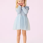 A young girl with blonde pigtails smiles against a pale pink background, wearing the Isobella and Chloe Tutu in the Clouds Dress, with its light blue tulle skirt and long-sleeved ribbed bodice, her hands gently touching her cheeks.