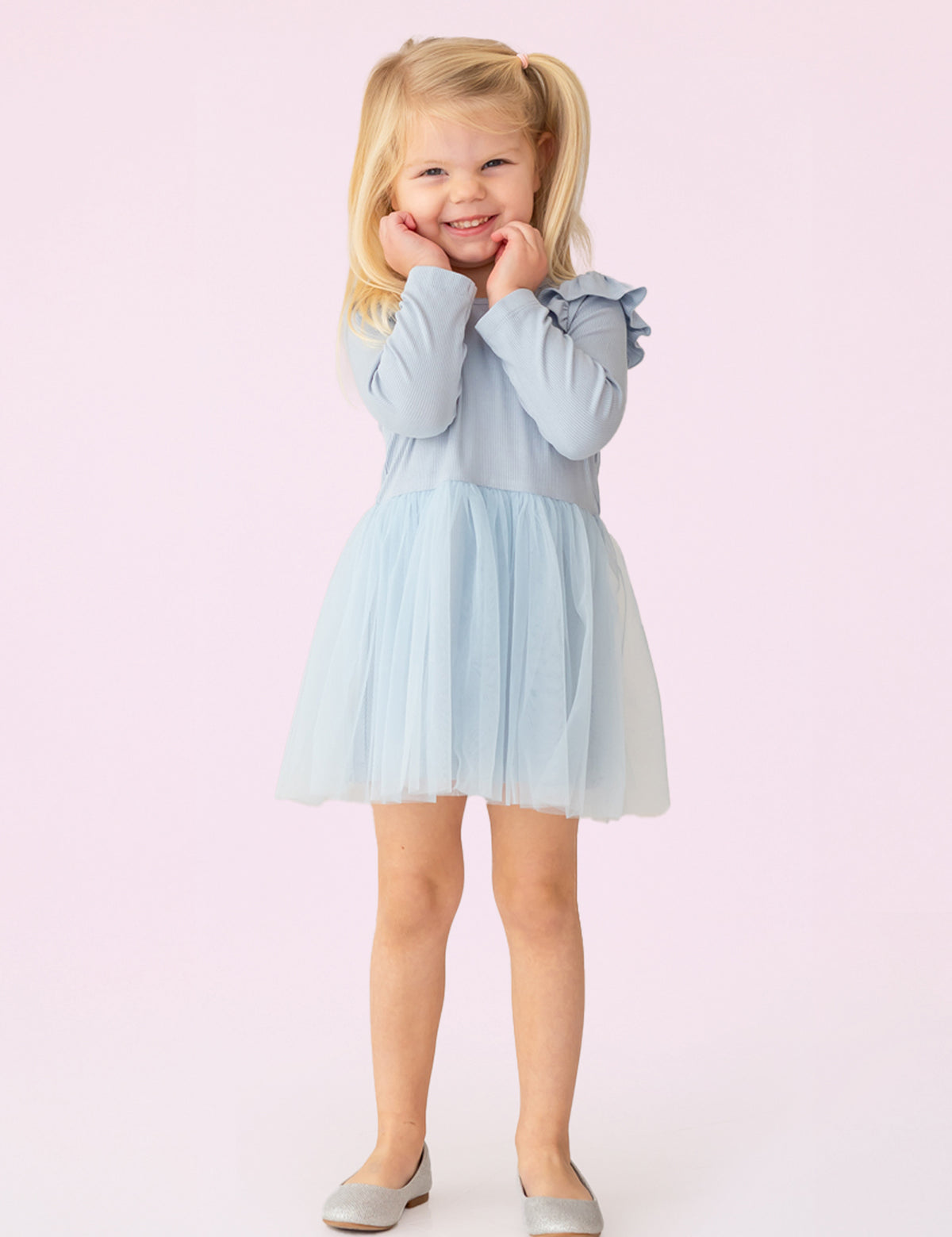 A young girl with blonde pigtails smiles against a pale pink background, wearing the Isobella and Chloe Tutu in the Clouds Dress, with its light blue tulle skirt and long-sleeved ribbed bodice, her hands gently touching her cheeks.