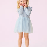 A young girl with blonde pigtails smiles against a pale pink background, wearing the Isobella and Chloe Tutu in the Clouds Dress, with its light blue tulle skirt and long-sleeved ribbed bodice, her hands gently touching her cheeks.