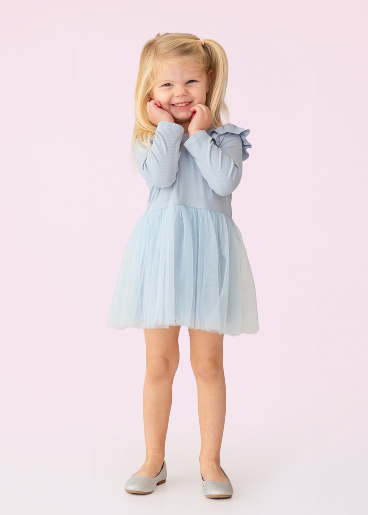 A young girl with blonde pigtails smiles against a pale pink background, wearing the Isobella and Chloe Tutu in the Clouds Dress, with its light blue tulle skirt and long-sleeved ribbed bodice, her hands gently touching her cheeks.