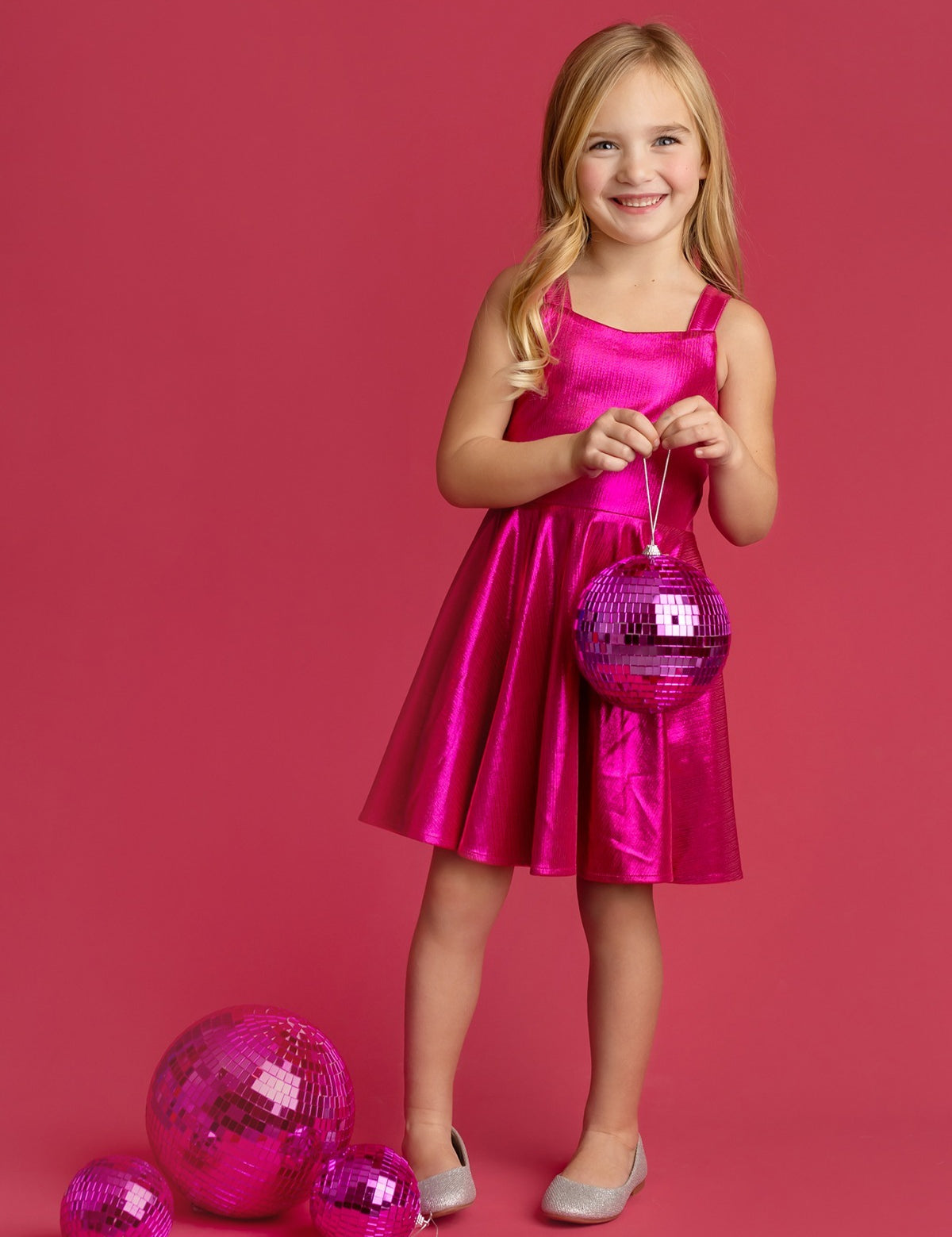 A young girl wears the Isobella and Chloe Glitterball Shimmering Sleeveless Dress, smiling against a pink background while holding a pink disco ball, with more disco balls at her feet. She completes the look with silver shoes.