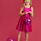 A young girl wears the Isobella and Chloe Glitterball Shimmering Sleeveless Dress, smiling against a pink background while holding a pink disco ball, with more disco balls at her feet. She completes the look with silver shoes.