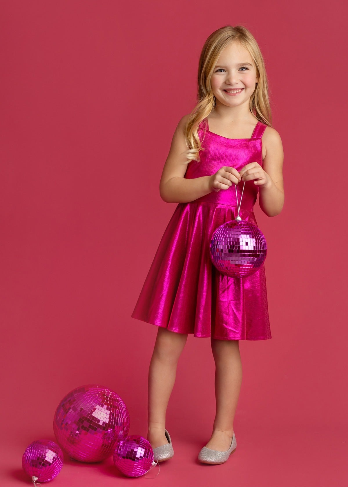 A young girl wears the Isobella and Chloe Glitterball Shimmering Sleeveless Dress, smiling against a pink background while holding a pink disco ball, with more disco balls at her feet. She completes the look with silver shoes.