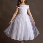 A young girl wears the Lauren Marie Satin Beaded Lace Bodice Dress, featuring a satin bodice and tulle skirt. She stands against a brown background, holding her skirt out, with wavy brown hair and light pink shoes.