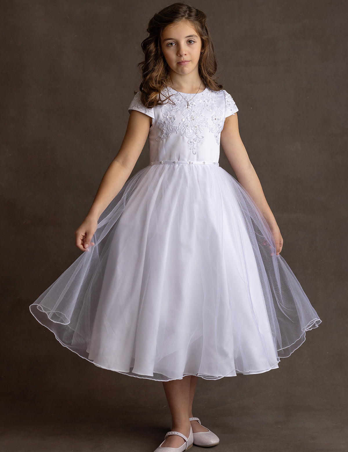 A young girl wears the Lauren Marie Satin Beaded Lace Bodice Dress, featuring a satin bodice and tulle skirt. She stands against a brown background, holding her skirt out, with wavy brown hair and light pink shoes.