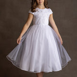 A young girl wears the Lauren Marie Satin Beaded Lace Bodice Dress, featuring a satin bodice and tulle skirt. She stands against a brown background, holding her skirt out, with wavy brown hair and light pink shoes.
