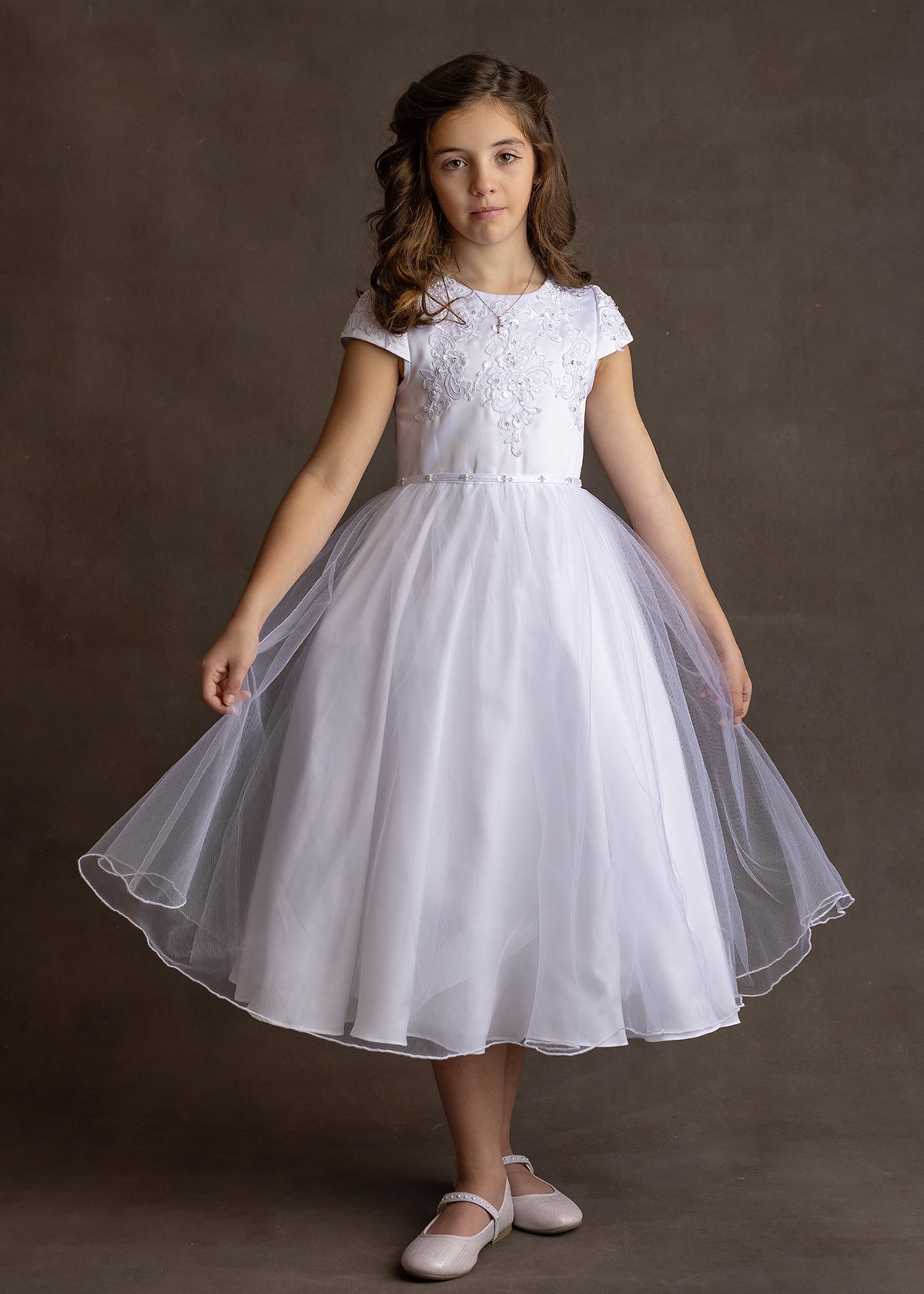 A young girl wears the Lauren Marie Satin Beaded Lace Bodice Dress, featuring a satin bodice and tulle skirt. She stands against a brown background, holding her skirt out, with wavy brown hair and light pink shoes.