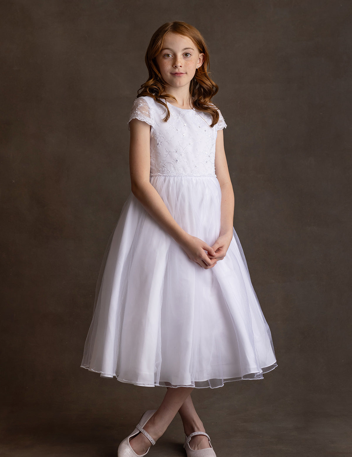 A young girl with red hair stands against a brown backdrop, wearing the Lauren Marie Scalloped Detail on Waist and Sleeve Dress, featuring a lace bodice, organza tulle skirt, and light pink shoes, hands clasped as she gazes at the camera.