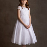 A young girl with red hair stands against a brown backdrop, wearing the Lauren Marie Scalloped Detail on Waist and Sleeve Dress, featuring a lace bodice, organza tulle skirt, and light pink shoes, hands clasped as she gazes at the camera.