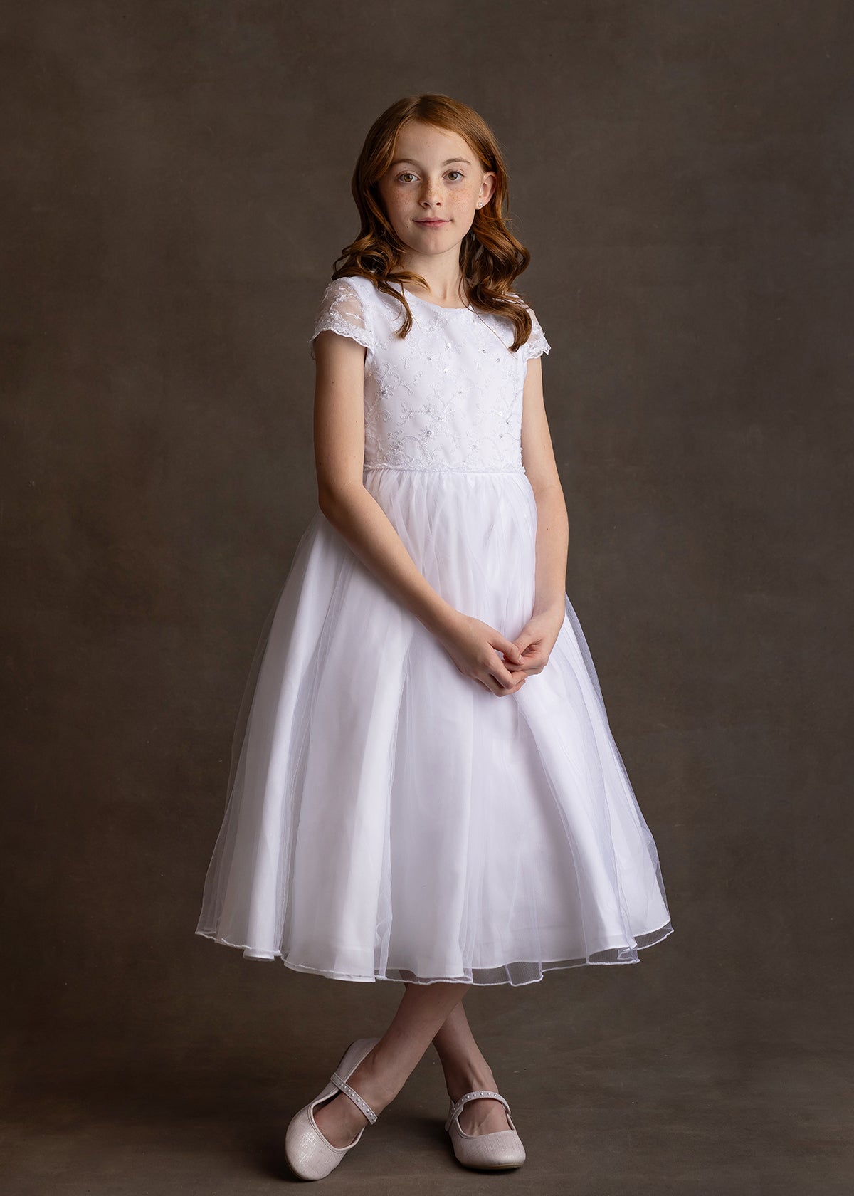 A young girl with red hair stands against a brown backdrop, wearing the Lauren Marie Scalloped Detail on Waist and Sleeve Dress, featuring a lace bodice, organza tulle skirt, and light pink shoes, hands clasped as she gazes at the camera.