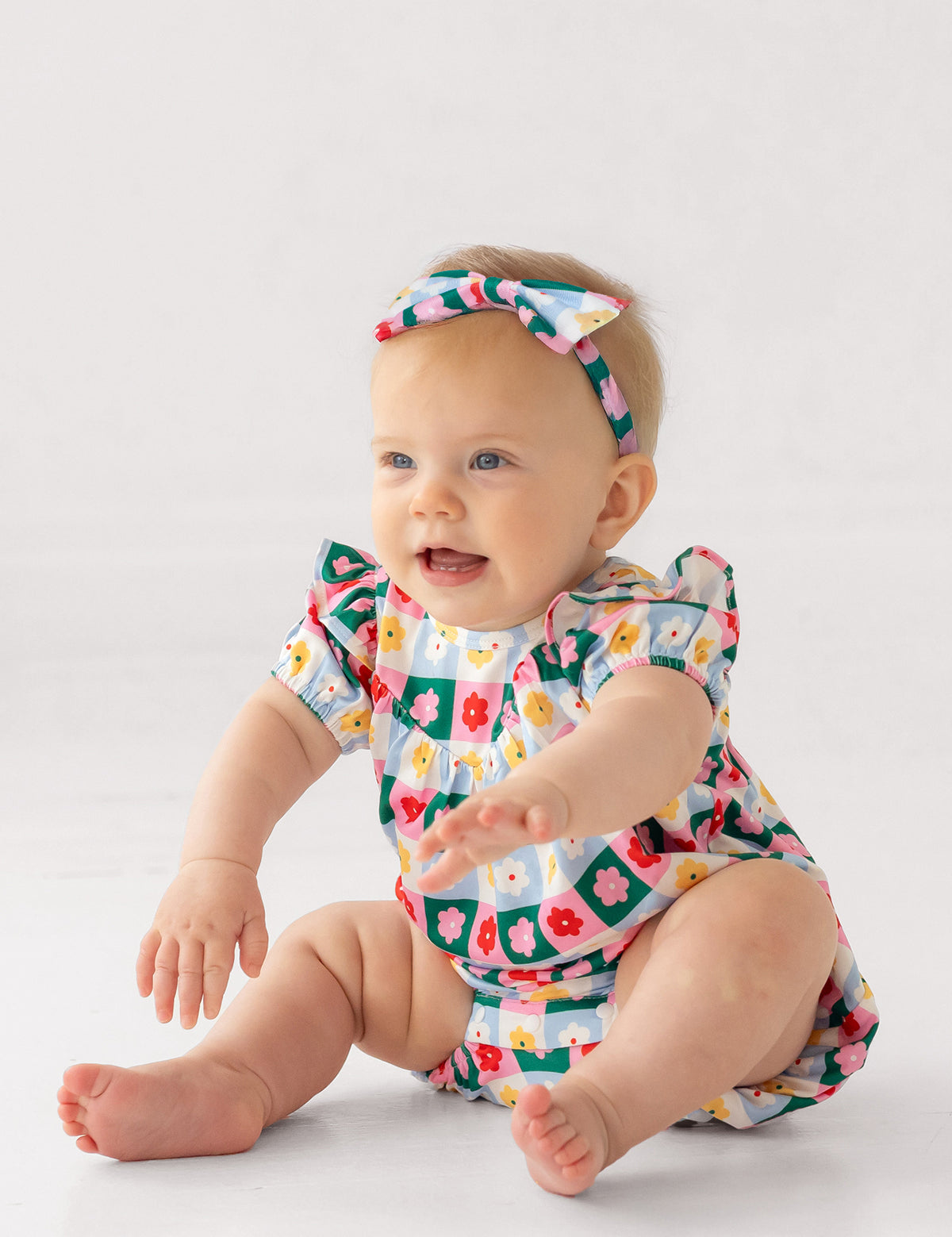 A smiling baby wearing the Mabel and Honey Flower Pops Short Sleeve Romper with a matching headband sits on a white floor, arms outstretched.