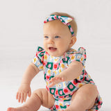 A smiling baby wearing the Mabel and Honey Flower Pops Short Sleeve Romper with a matching headband sits on a white floor, arms outstretched.