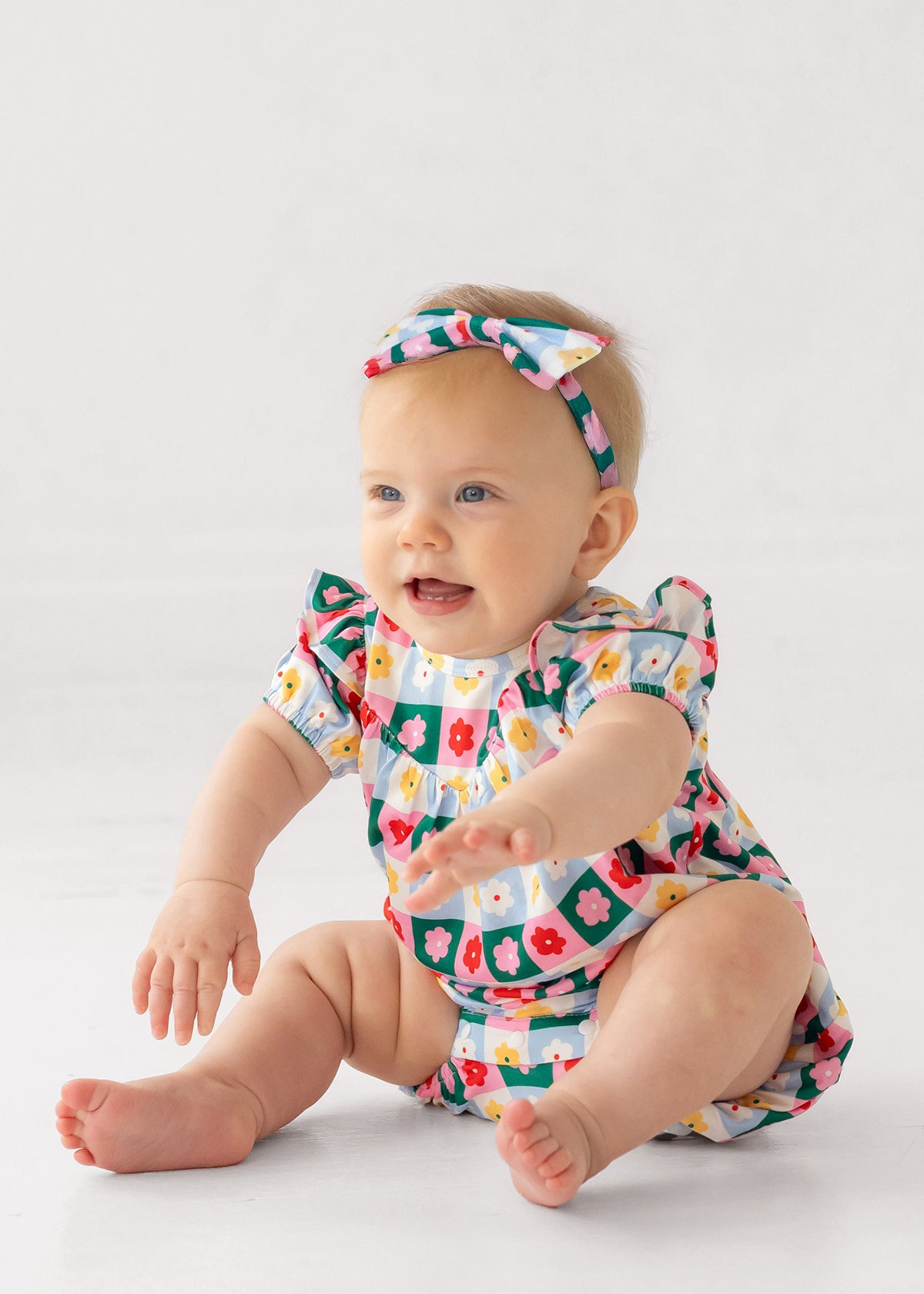 A smiling baby wearing the Mabel and Honey Flower Pops Short Sleeve Romper with a matching headband sits on a white floor, arms outstretched.