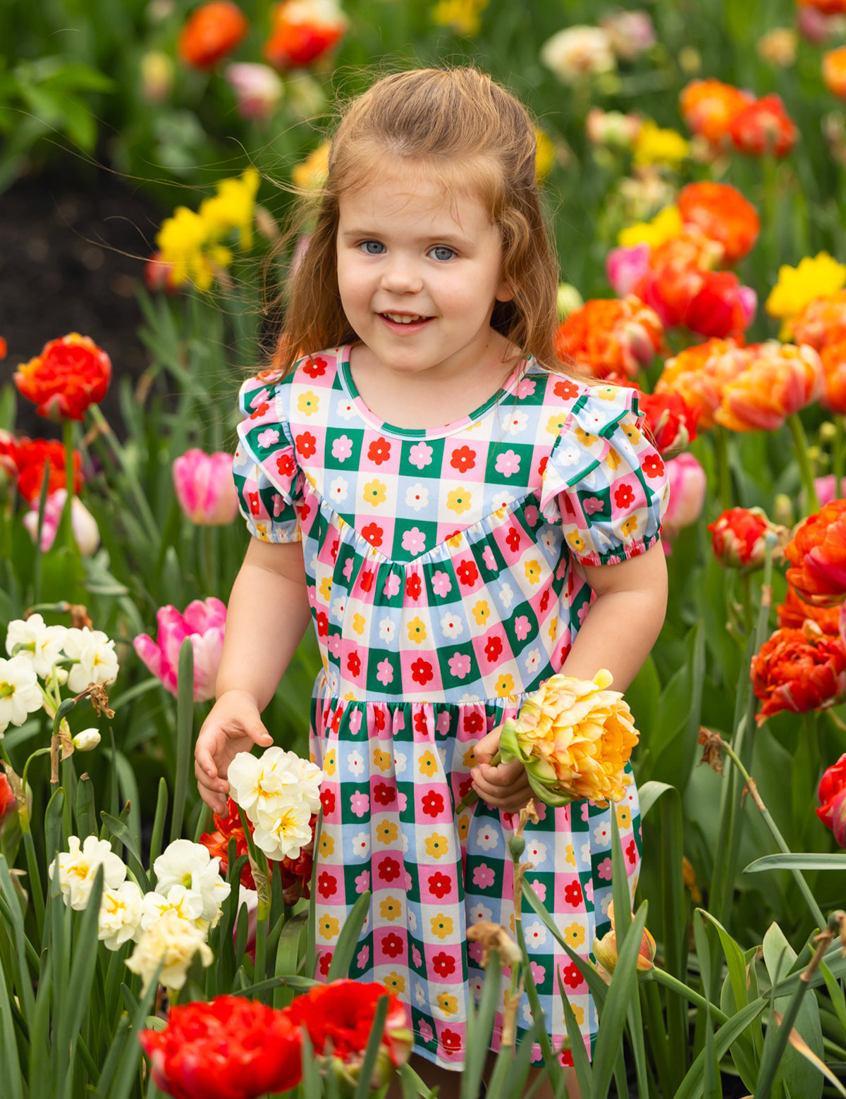 A young girl wears the vibrant Flower Pops Short Sleeve Dress by Mabel and Honey, standing among red, orange, yellow, and white flowers while holding a bouquet and smiling at the camera on a bright day.