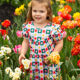 A young girl wears the vibrant Flower Pops Short Sleeve Dress by Mabel and Honey, standing among red, orange, yellow, and white flowers while holding a bouquet and smiling at the camera on a bright day.